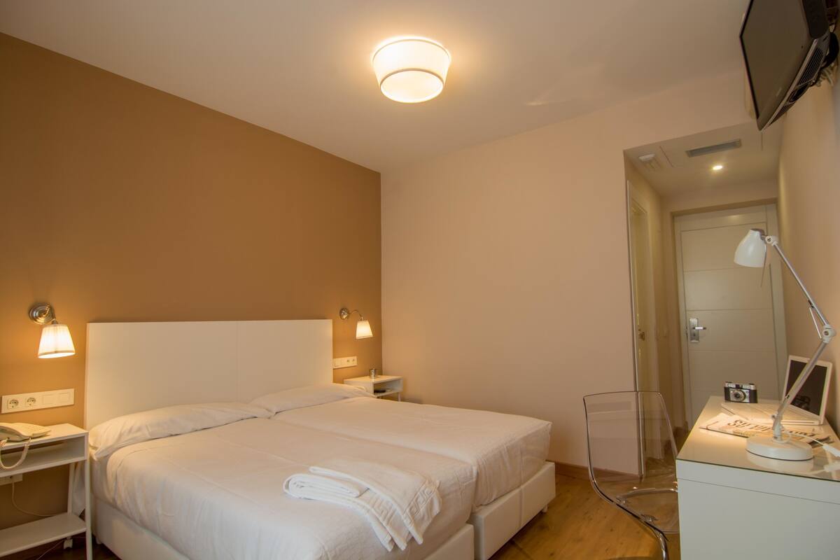 Una habitación de hotel con cama, lámparas de noche, un escritorio con lámpara y un espejo.