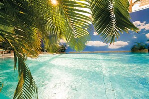 Une piscine d’un bleu éclatant, bordée de palmiers, sous un ciel dégagé et avec une plage qui s’étend au loin.