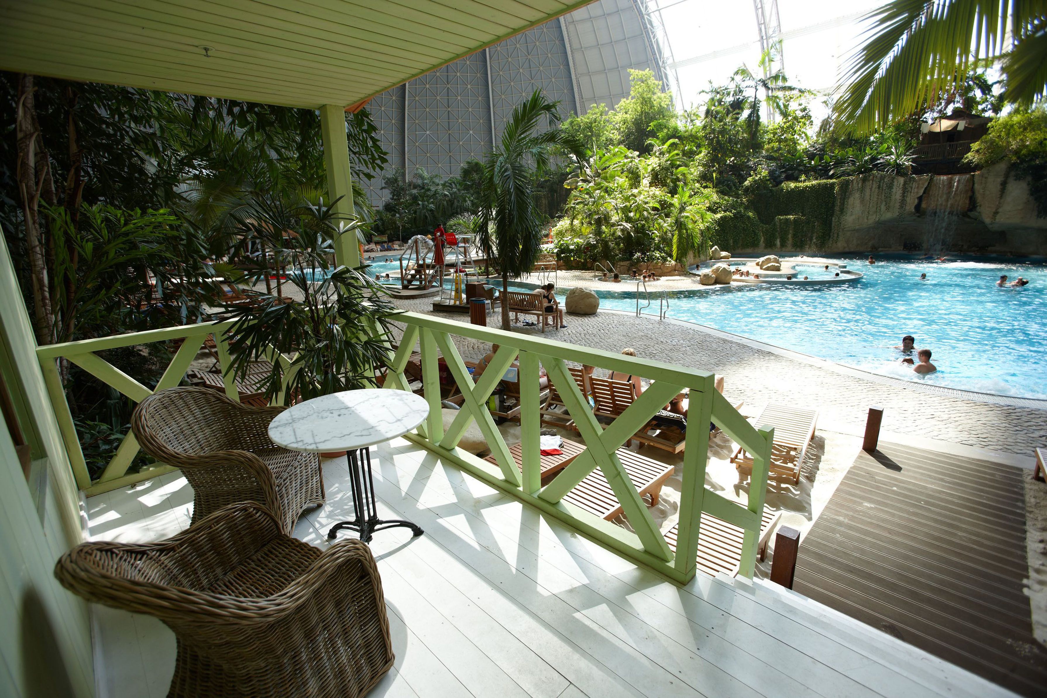 A pool area with lounge chairs and tables, surrounded by tropical plants and a large building in the background.