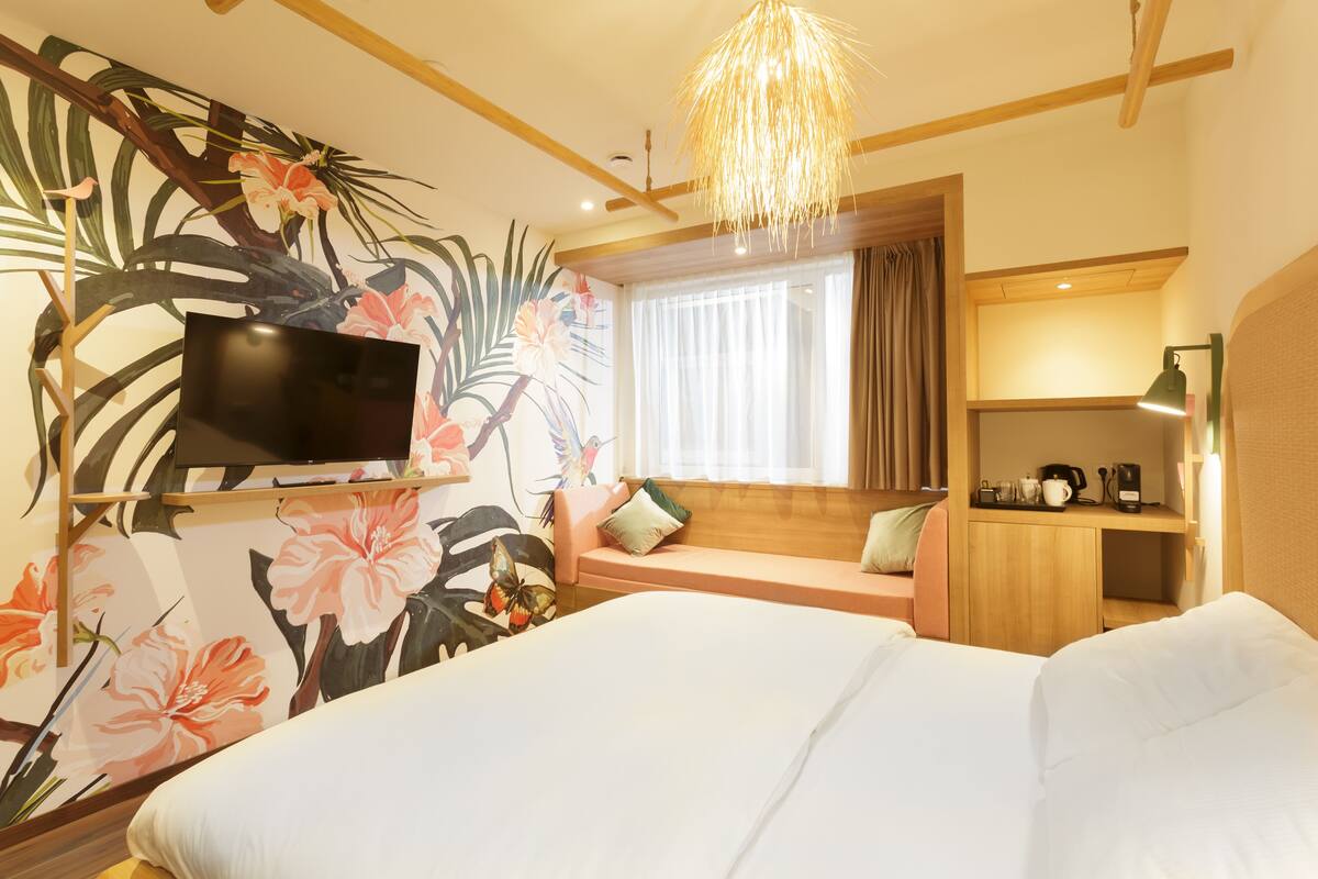 Une chambre d’hôtel comprenant un lit, un téléviseur fixé au mur, un coin salon avec des coussins, un bureau avec une lampe et une fenêtre avec des rideaux.