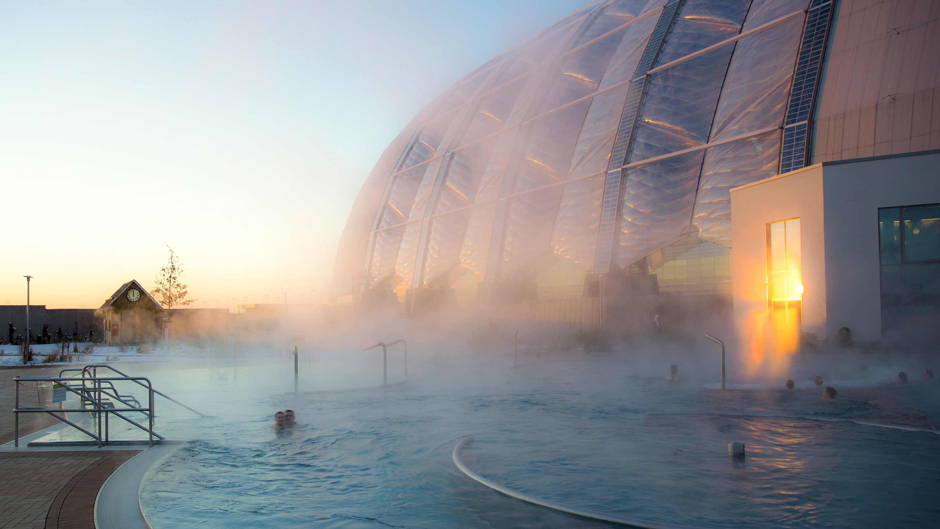 A geothermal spa with a large pool, steam rising from the water, and a modern building with a glass facade in the background.