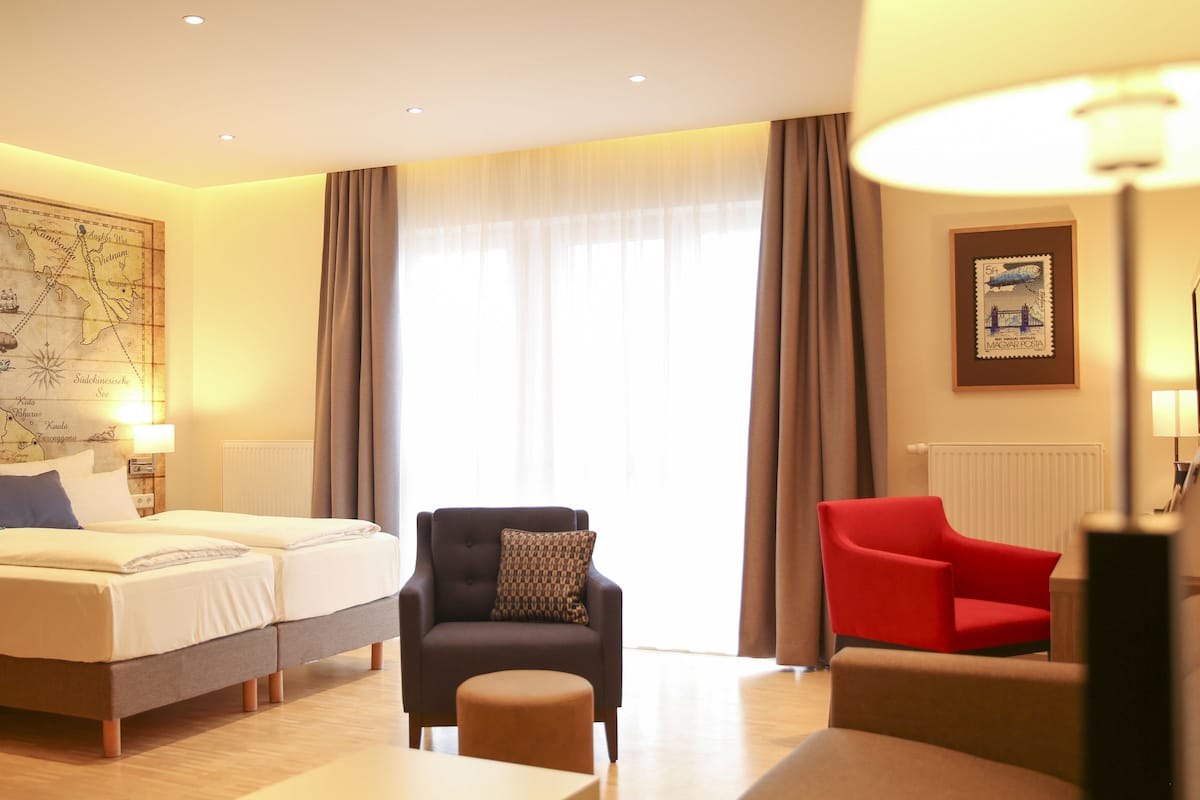 Une chambre d’hôtel avec un grand lit, deux tables de chevet, une carte accrochée au mur, un fauteuil rouge, un fauteuil marron et une fenêtre avec des rideaux.