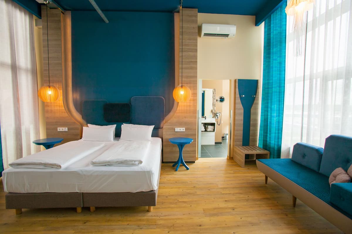 Une chambre d’hôtel moderne avec un grand lit, un canapé et une petite table. On aperçoit une salle de bain au fond.
