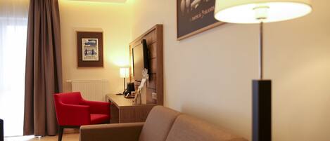A hotel room with a sofa, a red chair, a desk, a lamp, a radiator, and a framed picture on the wall.