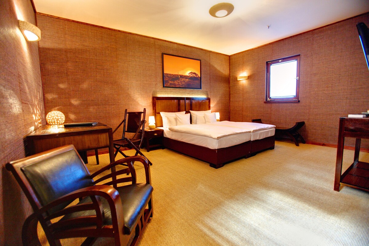 Une chambre d’hôtel avec un grand lit, un bureau avec une lampe, deux chaises, une fenêtre et un tableau au mur.