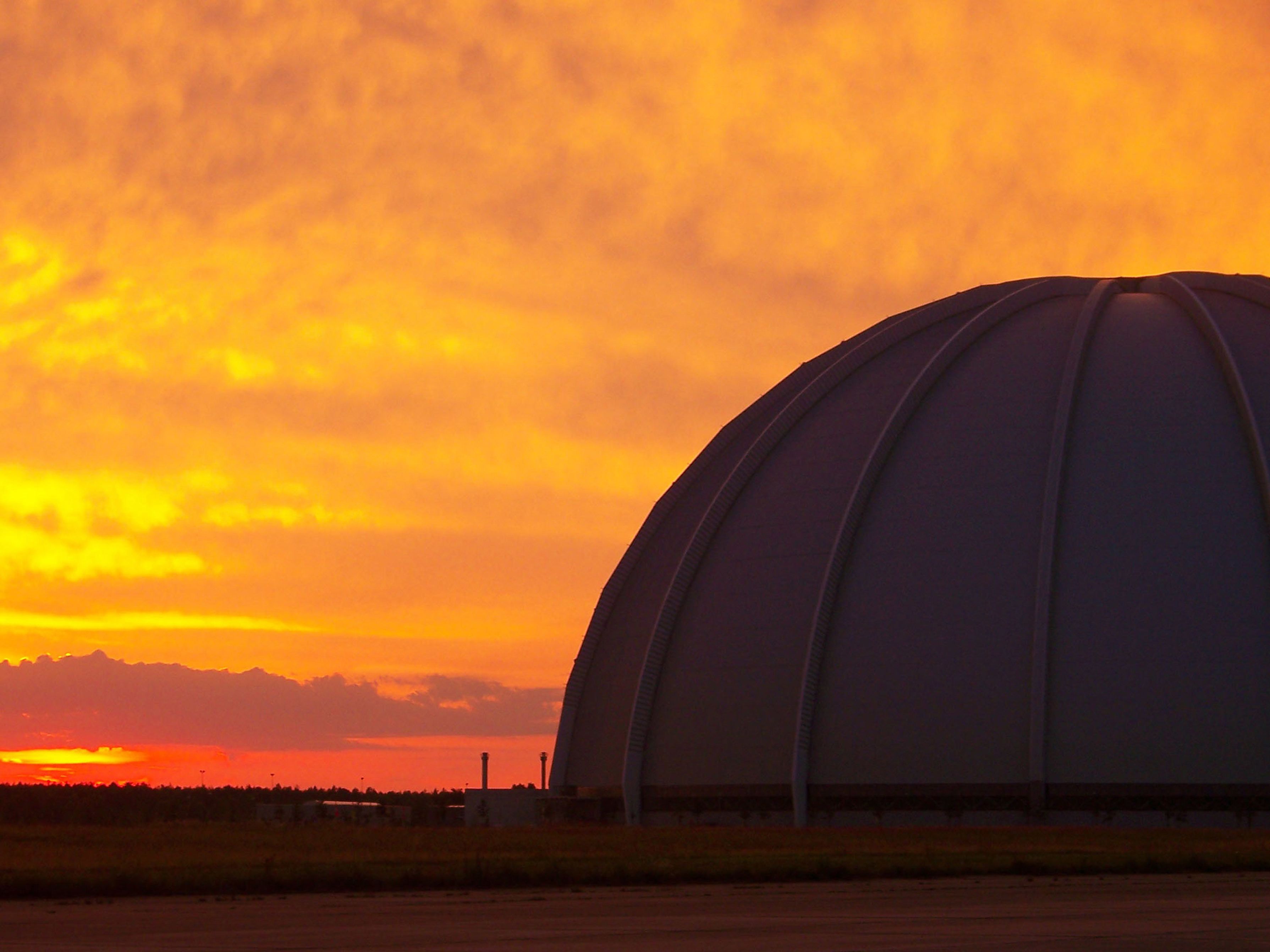 Ein großes, kuppelartiges Gebäude mit flachem Dach, eingebettet in eine flache Landschaft mit Sonnenuntergang im Hintergrund.