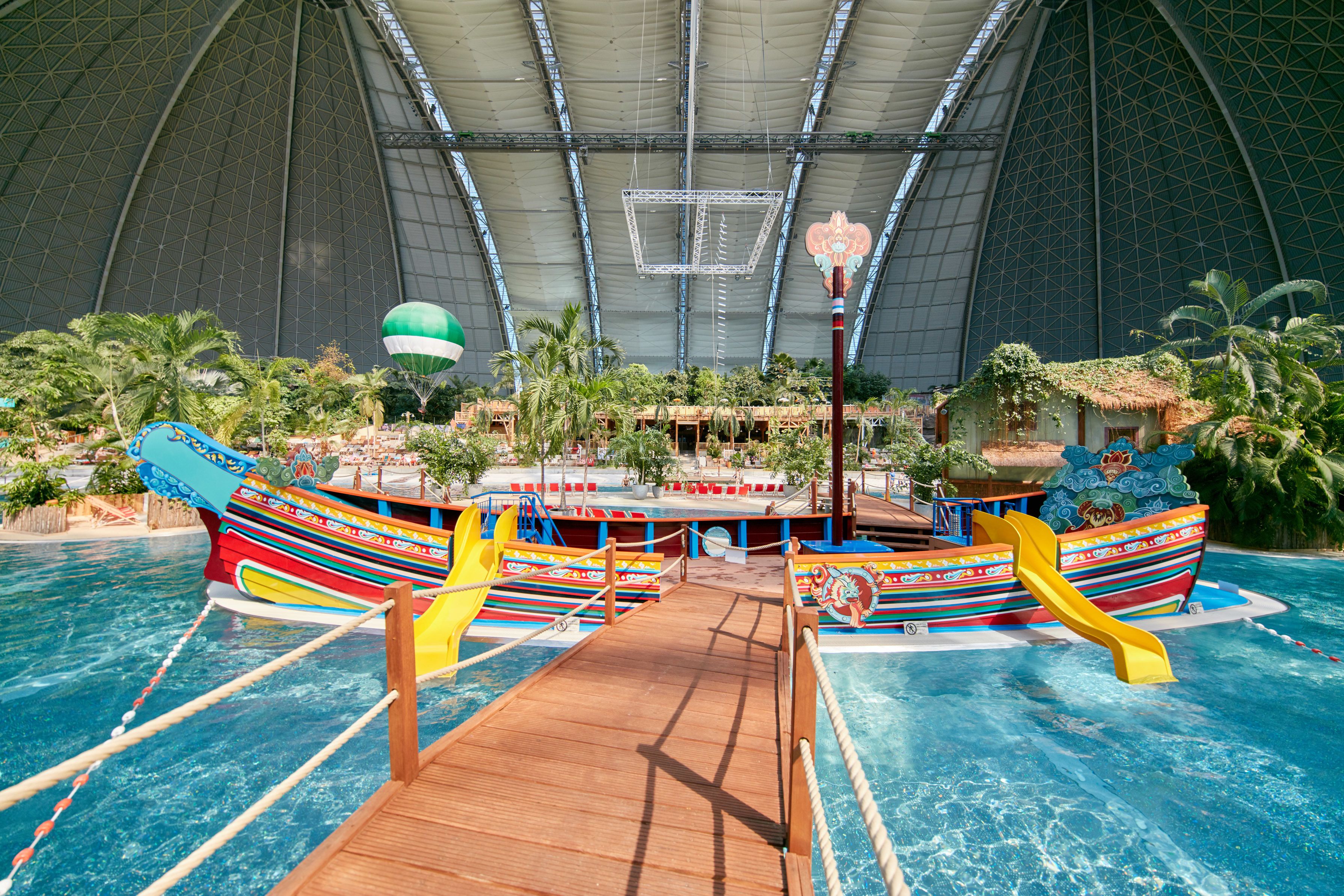 An indoor water park with a large pool, two colorful boat slides, and a wooden deck.