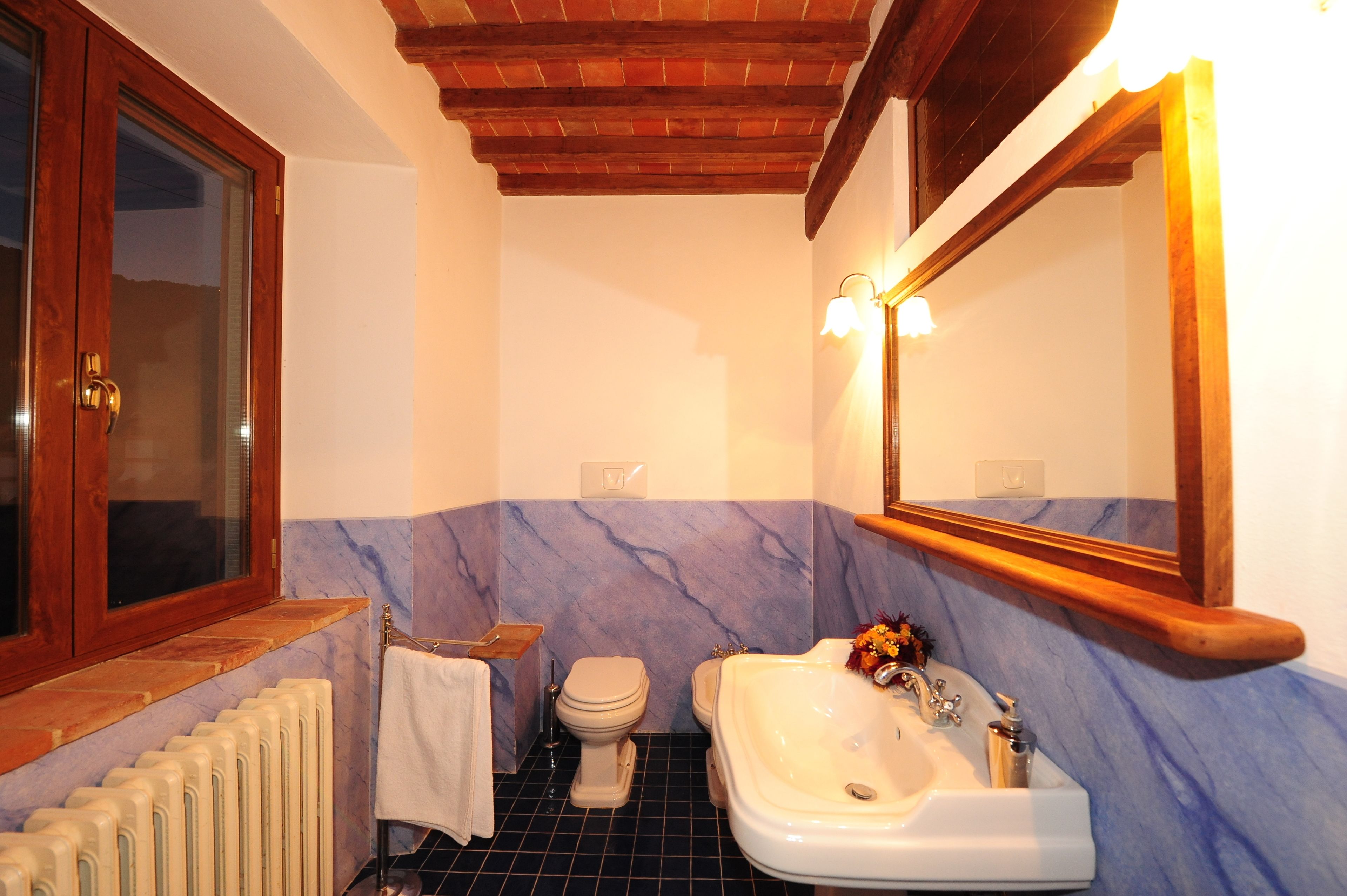 Ein Badezimmer mit Holzboden, Marmorwand, Waschbecken in Weiß, Toilette und einem Spiegel mit Beleuchtung.
