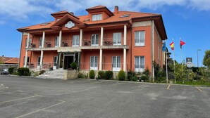 A two-story hotel with a red-tiled roof, a balcony on the second floor, and a parking lot in front. - Hotel Villa de Llanes (Llanes)