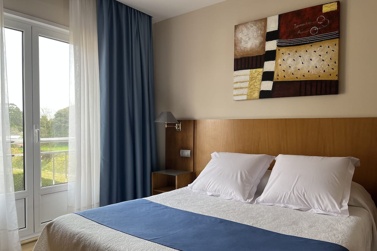Een hotelkamer met een bed, uitzicht door een raam, een schilderij aan de muur en een nachtkastje.