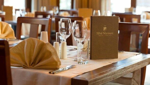 A dining table set with a menu, glasses, and cutlery in a well-lit room with wooden furniture. - Marmore (Cervinia)