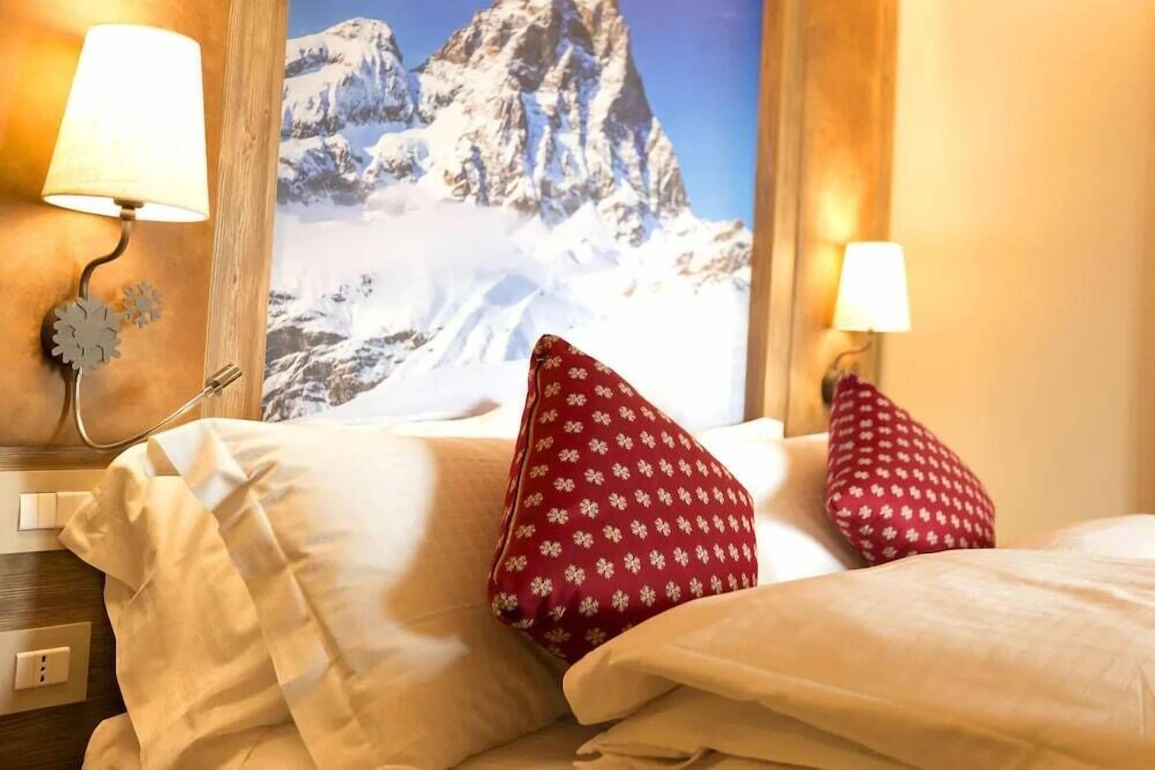 Quarto de hotel com uma cama, dois travesseiros vermelhos estampados, uma mesa de cabeceira com abajur e um candeeiro fixo na parede.