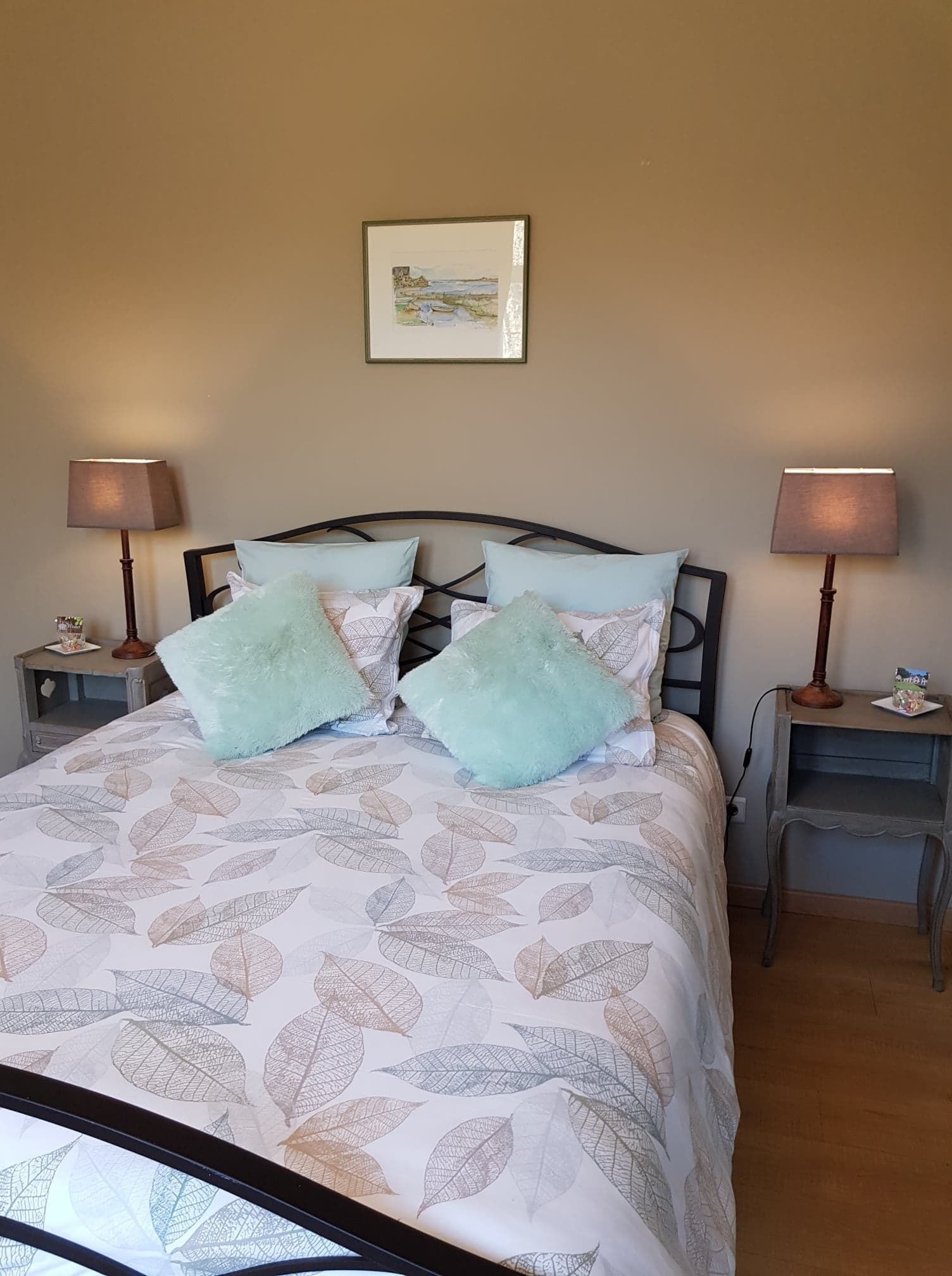 A neatly made bed with a leaf-patterned bedspread, two fluffy pillows, and a framed picture on the wall.