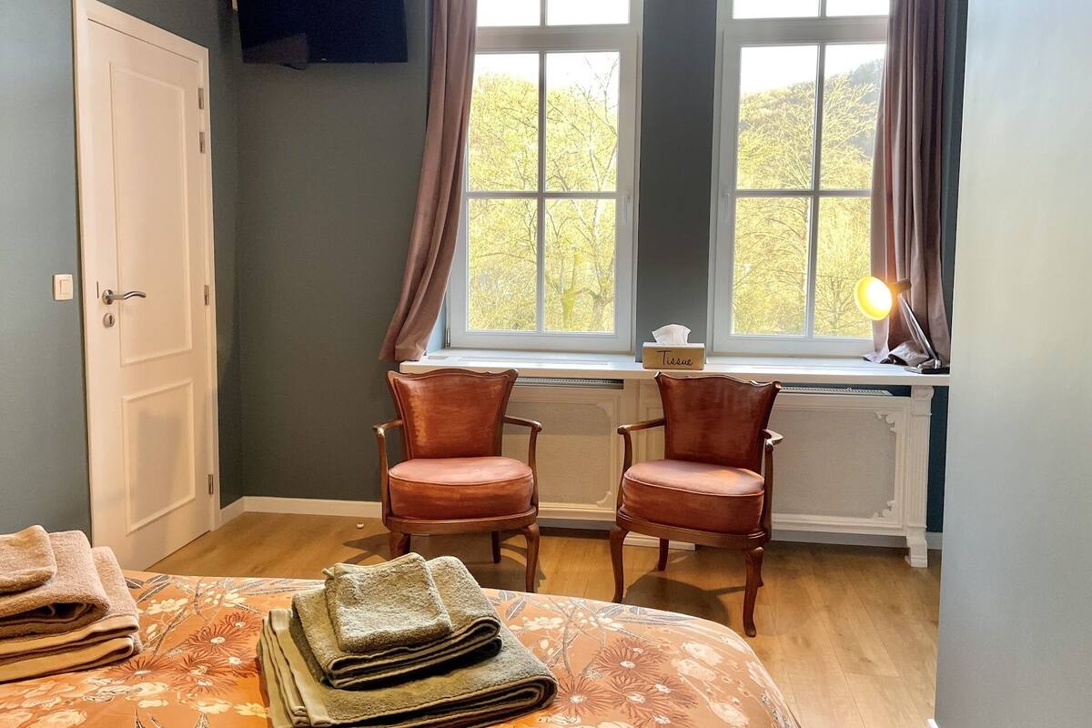 Une chambre à coucher avec deux chaises en bois, un lit recouvert d’une couvre-lit à motifs floraux, une fenêtre avec des rideaux et une table de chevet sur laquelle se trouvent une lampe et une boîte de mouchoirs.