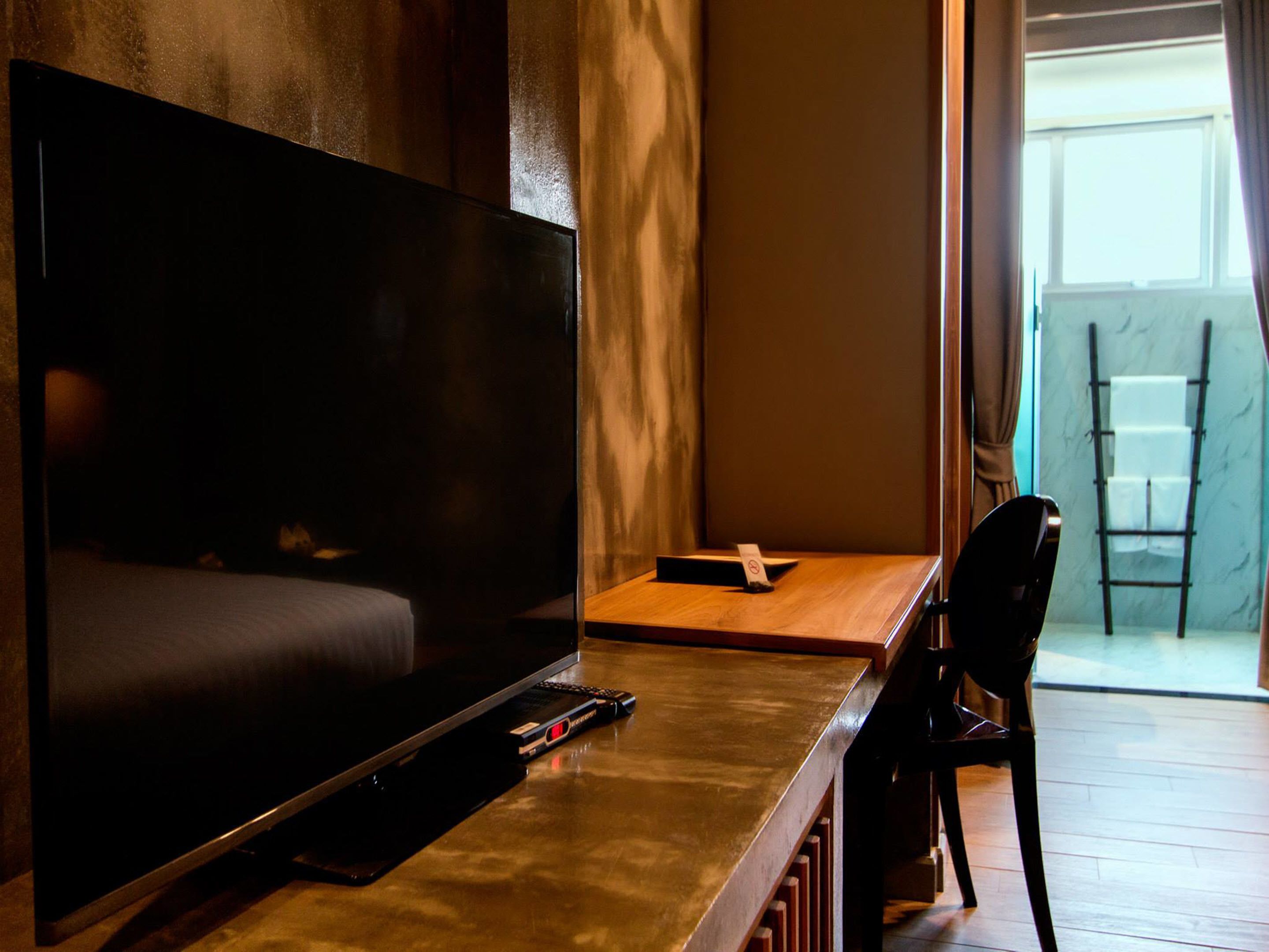Une chambre d’hôtel moderne avec un grand téléviseur à écran plat posé sur une table en bois, une télécommande et une petite chaise noire.