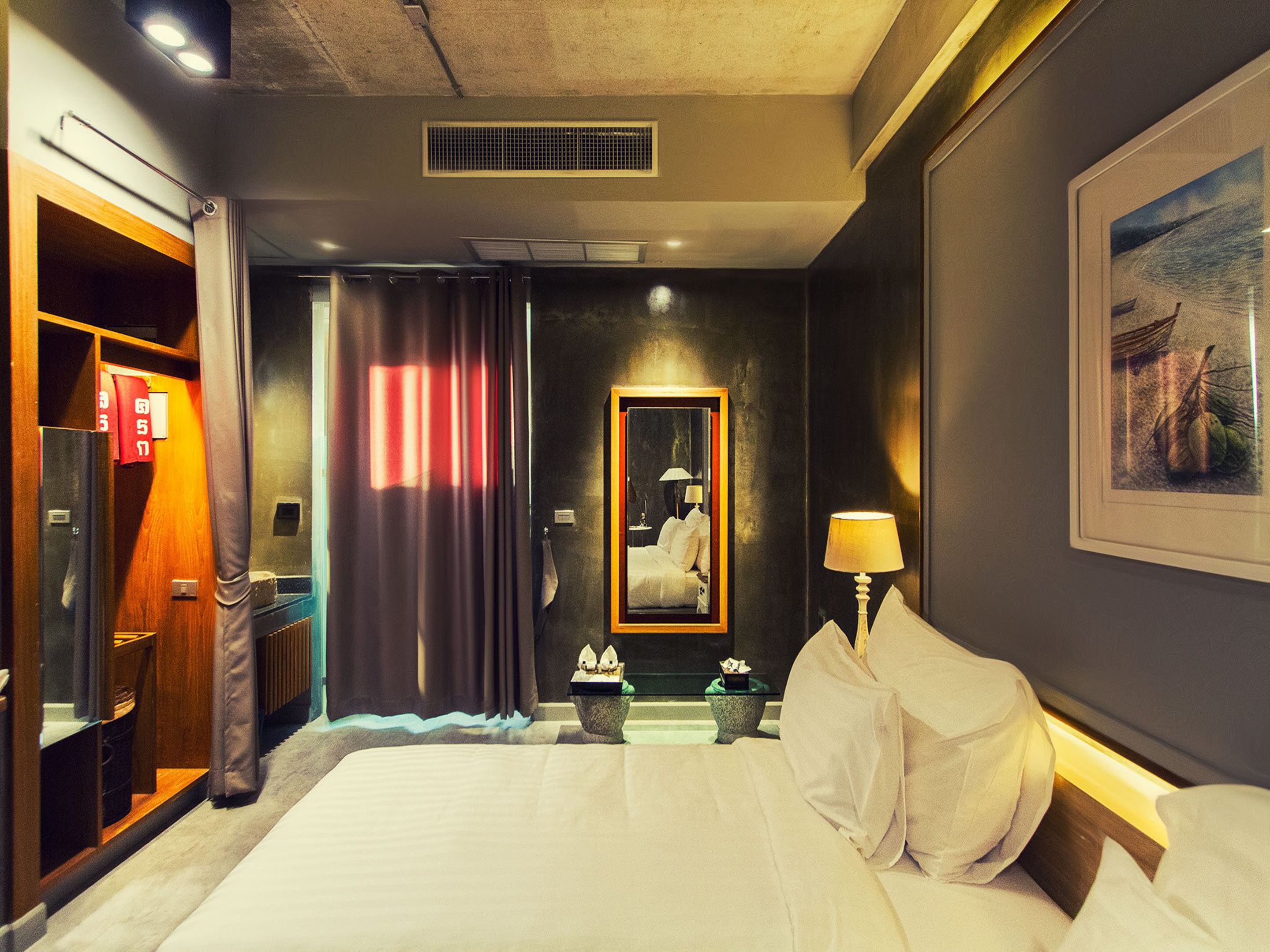 Quarto de hotel com uma cama grande, uma mesa de cabeceira com abajur, um espelho, um quadro na parede e uma janela com cortinas.