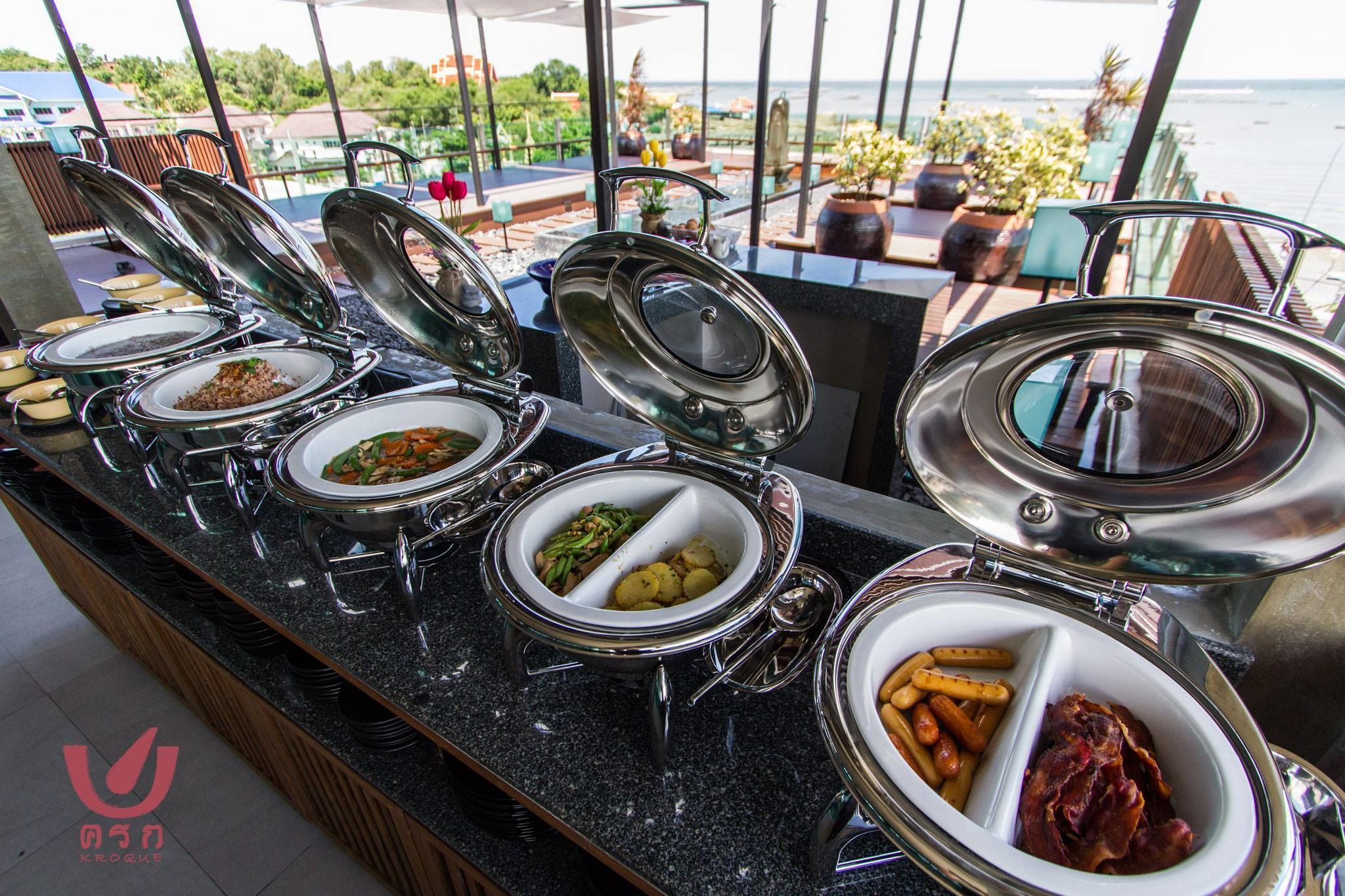 Montaje de bufé con diferentes alimentos en recipientes cubiertos, frente a un paisaje pintoresco.