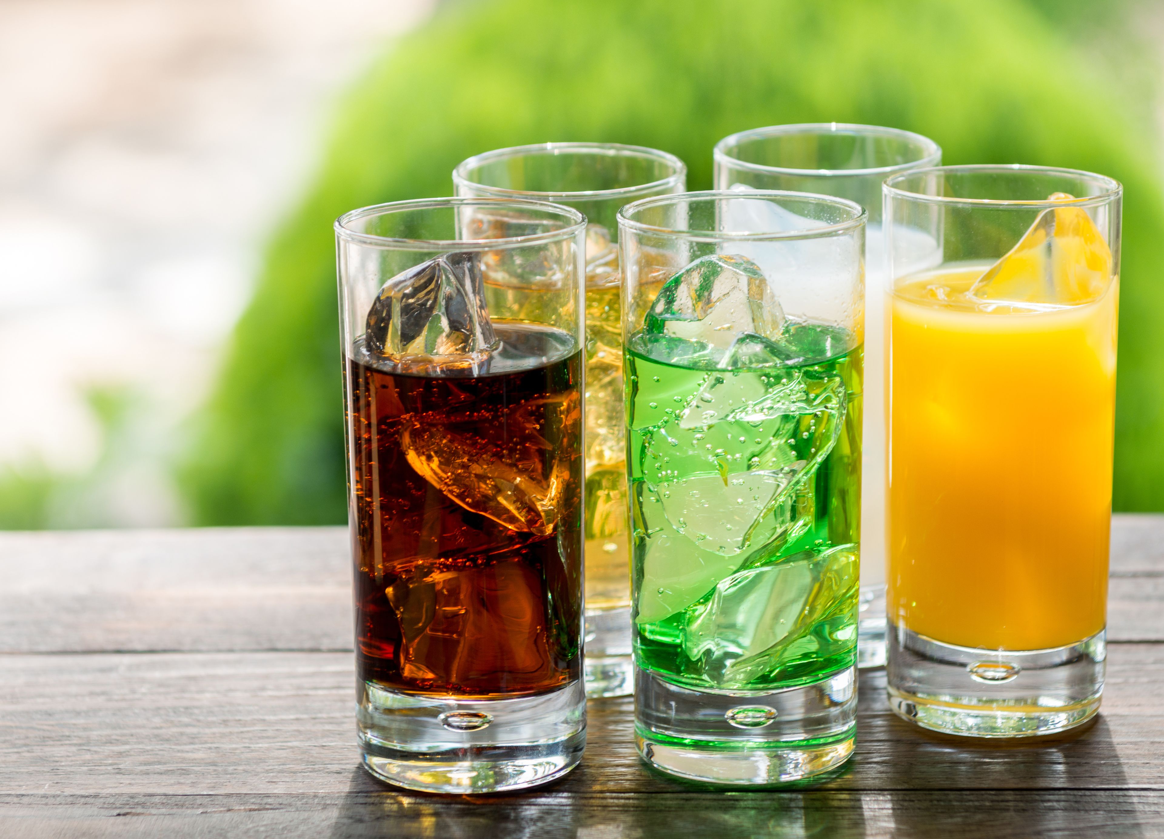 Four glasses of different colored beverages on a wooden table with a blurred green background.