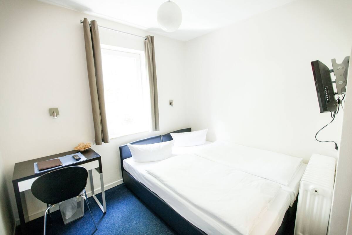 Habitación de hotel con cama, escritorio, silla, ventana con cortinas y televisor montado en la pared.