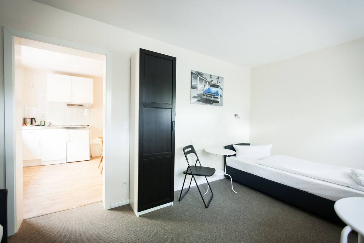Habitación de hotel moderna con una cama, una mesita y una silla. Hay una kitchenette en la habitación contigua.