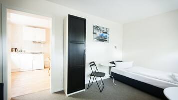 A modern hotel room with a bed, a small table, and a chair. There is a kitchenette in the adjacent room.