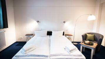 A hotel room with two beds, a chair, a table with a plate of food, and a lamp.
