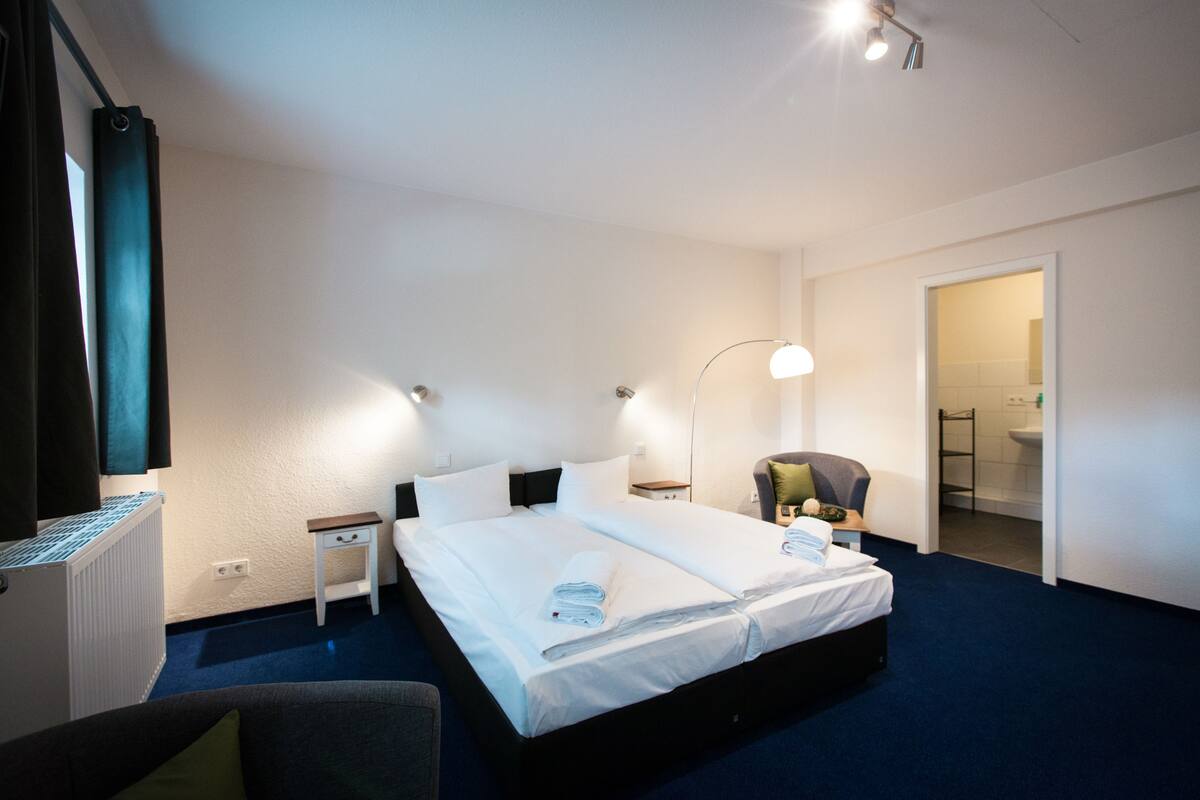 Habitación de hotel con una cama grande, mesitas de noche, una silla, un escritorio y un baño visible a través de una puerta abierta.
