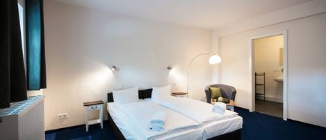 A hotel room with a large bed, bedside tables, a chair, a desk, and a bathroom visible through an open door.