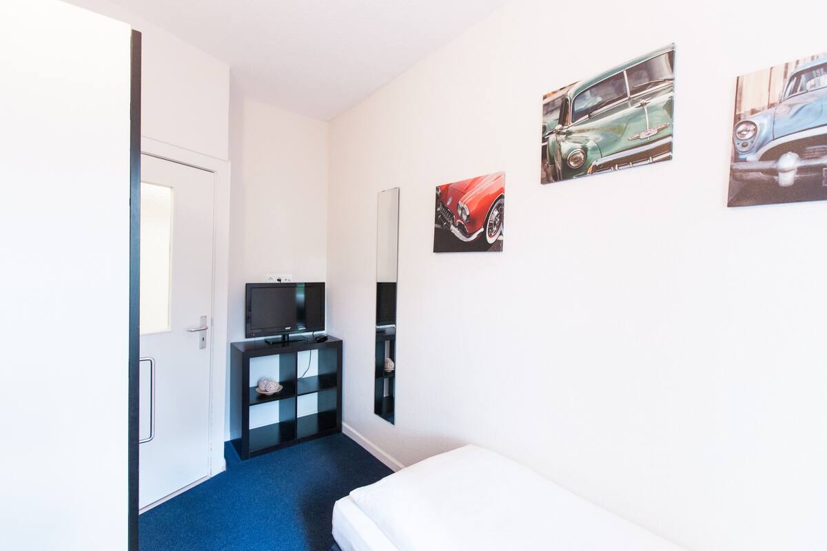 Una habitación de hotel con una cama, un televisor sobre un soporte negro, un estante pequeño con adornos y tres cuadros de autos antiguos colgados en la pared.