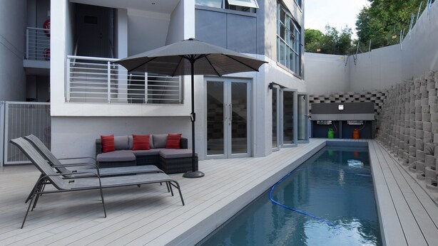 A modern outdoor pool area with a lounge chair and a table, a covered seating area, and a pool with a blue hose. - Insignia Lifestyle (Sandton)