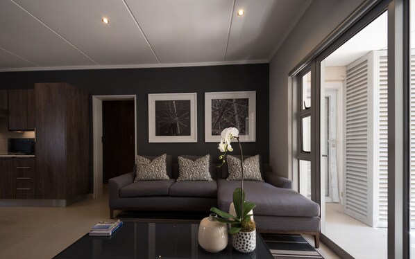 Una sala de estar moderna con un sofá gris, cojines estampados, una mesa de centro con libros y un jarrón, y arte abstracto enmarcados en la pared.