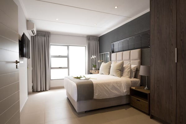 A modern bedroom with a large bed, a TV, a bedside table with a lamp, and a wardrobe.