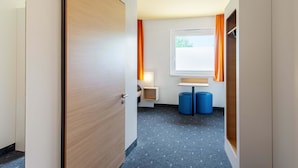 A hotel room with a wooden door, a window with orange curtains, a small desk, and two blue ottomans. - B&B Hotel Augsburg-Süd (Augsburg)