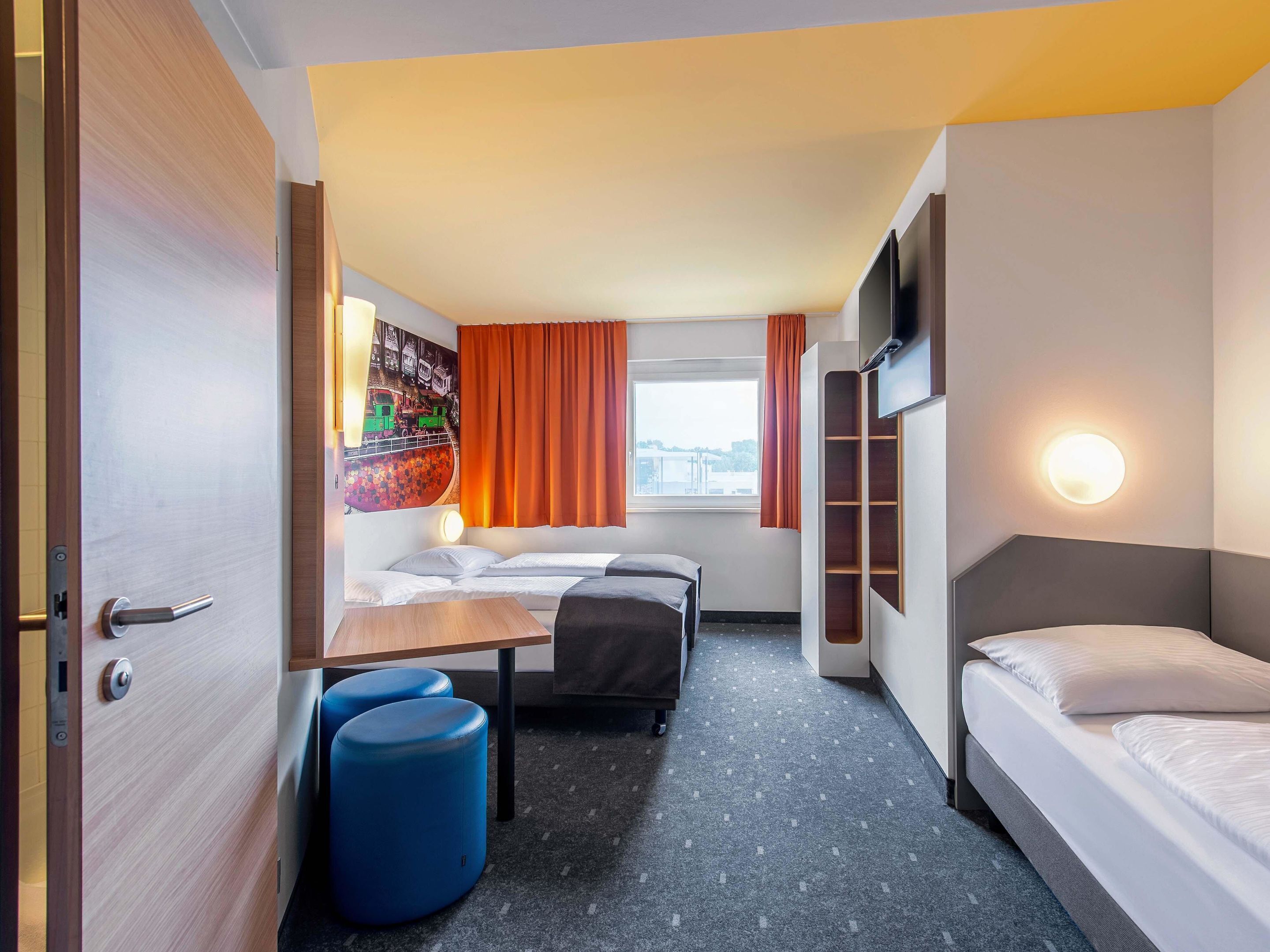 Una habitación de hotel con cama, escritorio, dos taburetes azules, televisor montado en la pared y ventana con cortinas.