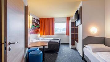 Una habitación de hotel con cama, escritorio, dos taburetes azules, televisor montado en la pared y ventana con cortinas.