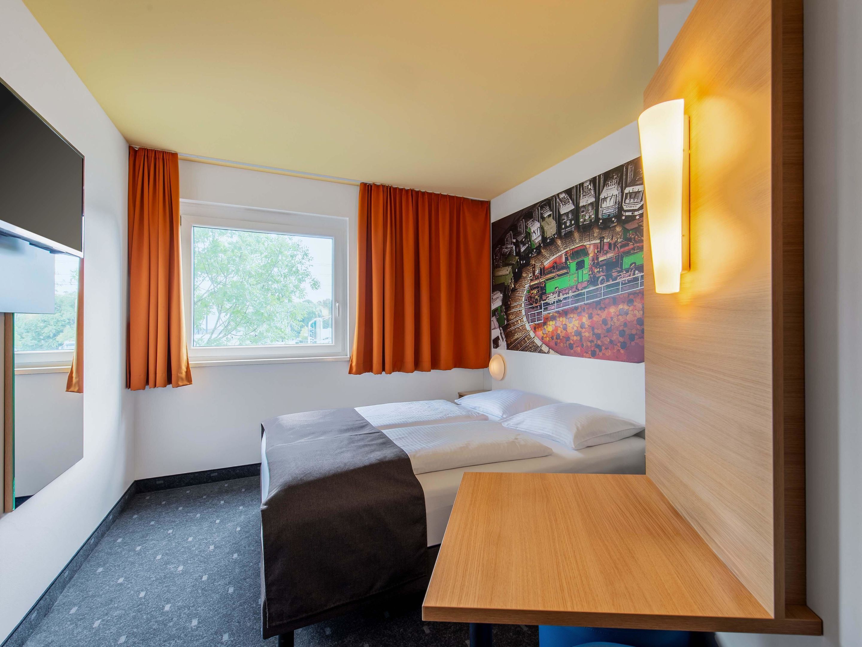 A hotel room with a bed, a small table, a wall-mounted light, a window with curtains, and a mural on the wall.
