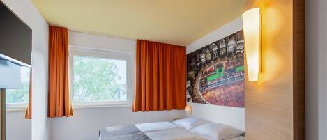 Habitación de hotel con cama, una mesita, luz de pared, ventana con cortinas y un mural en la pared.