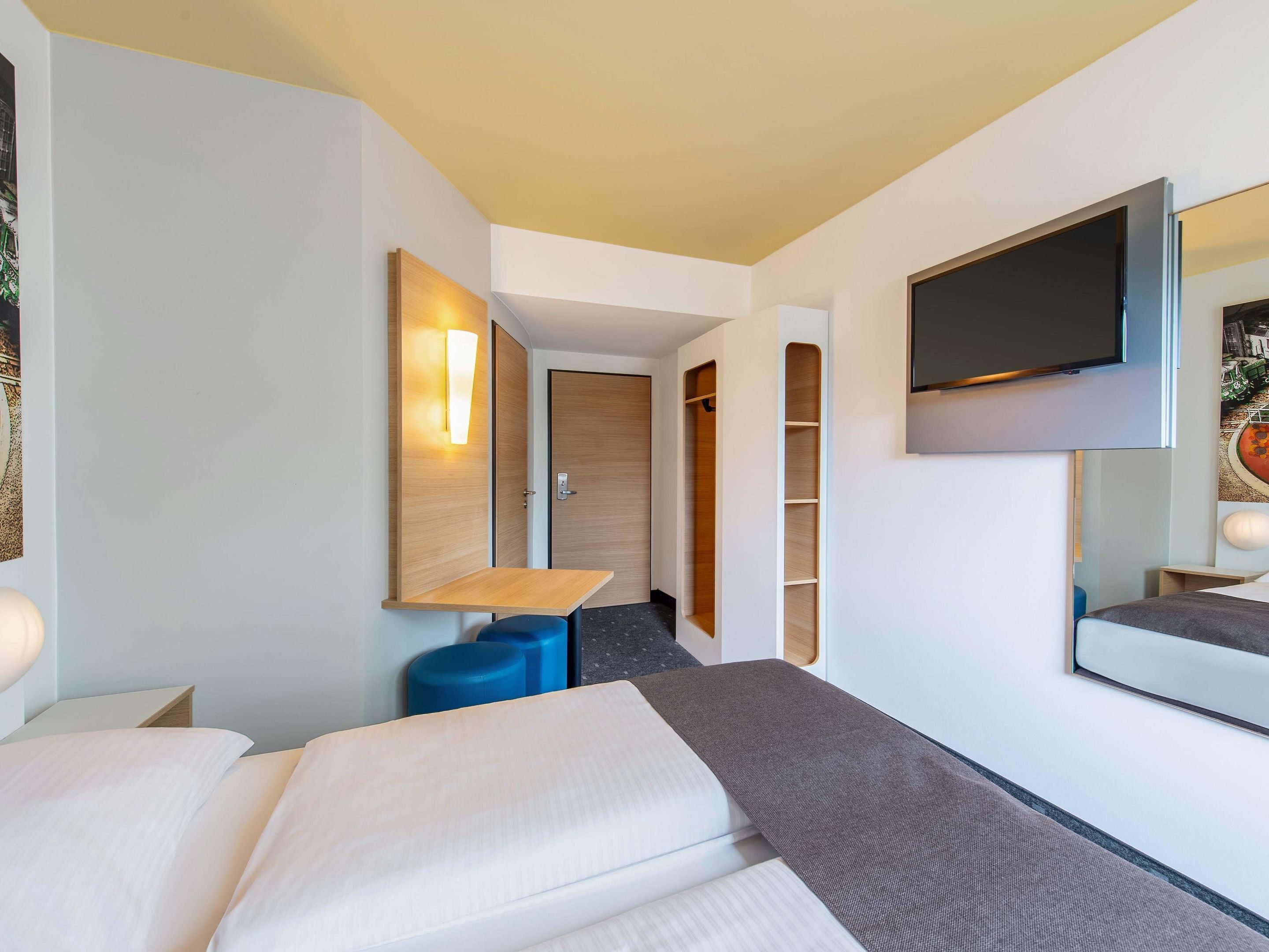 A modern hotel room with a bed, a desk, a TV, and a wardrobe.
