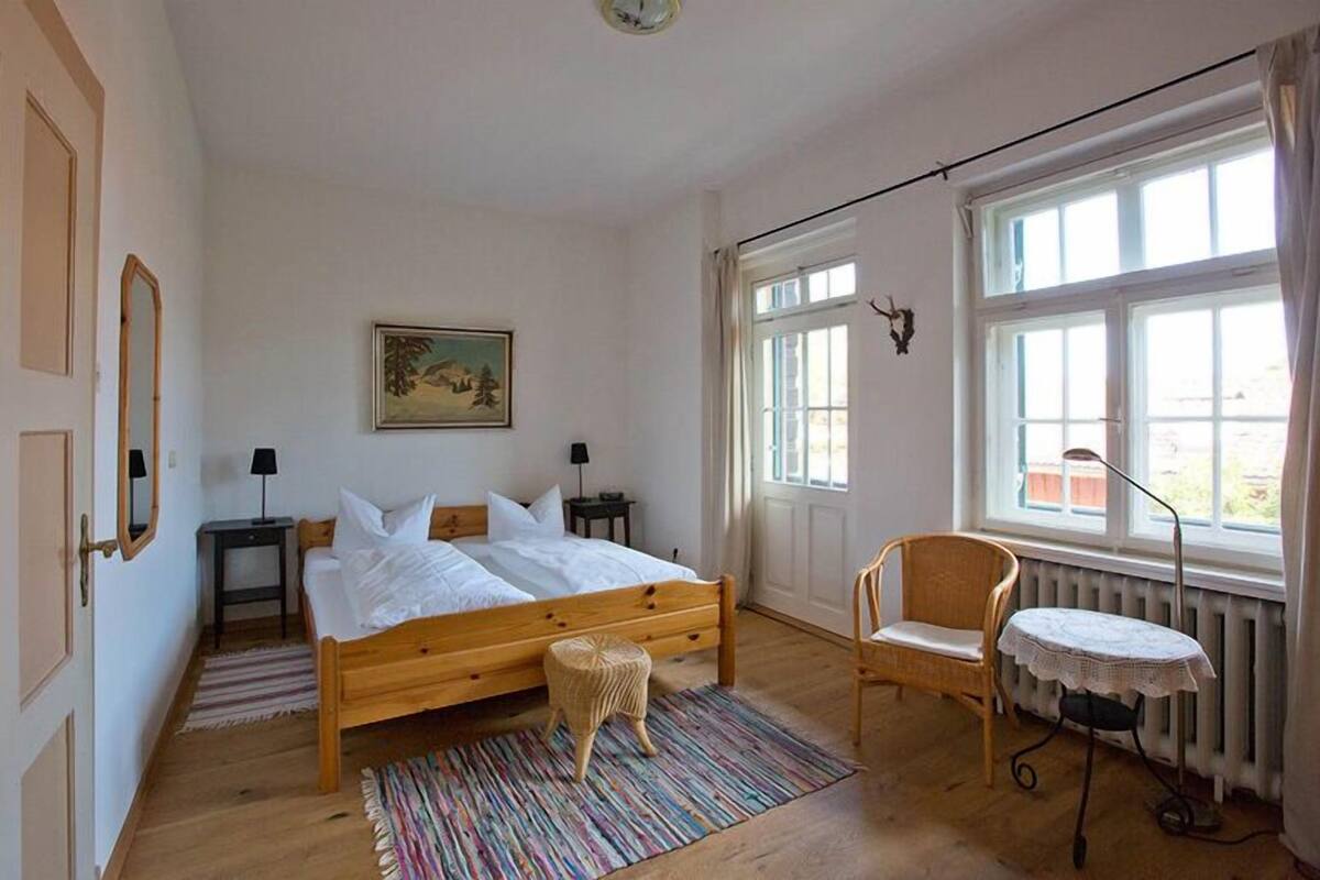 A bedroom with a wooden bed, a small round table, a chair, a radiator, and a window with curtains.