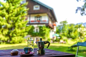 A table with a coffee pot, a glass of water, and a muffin on a wooden table in a garden setting with a building in the background. - Gästehaus Hohe Tannen (Garmisch-Partenkirchen)
