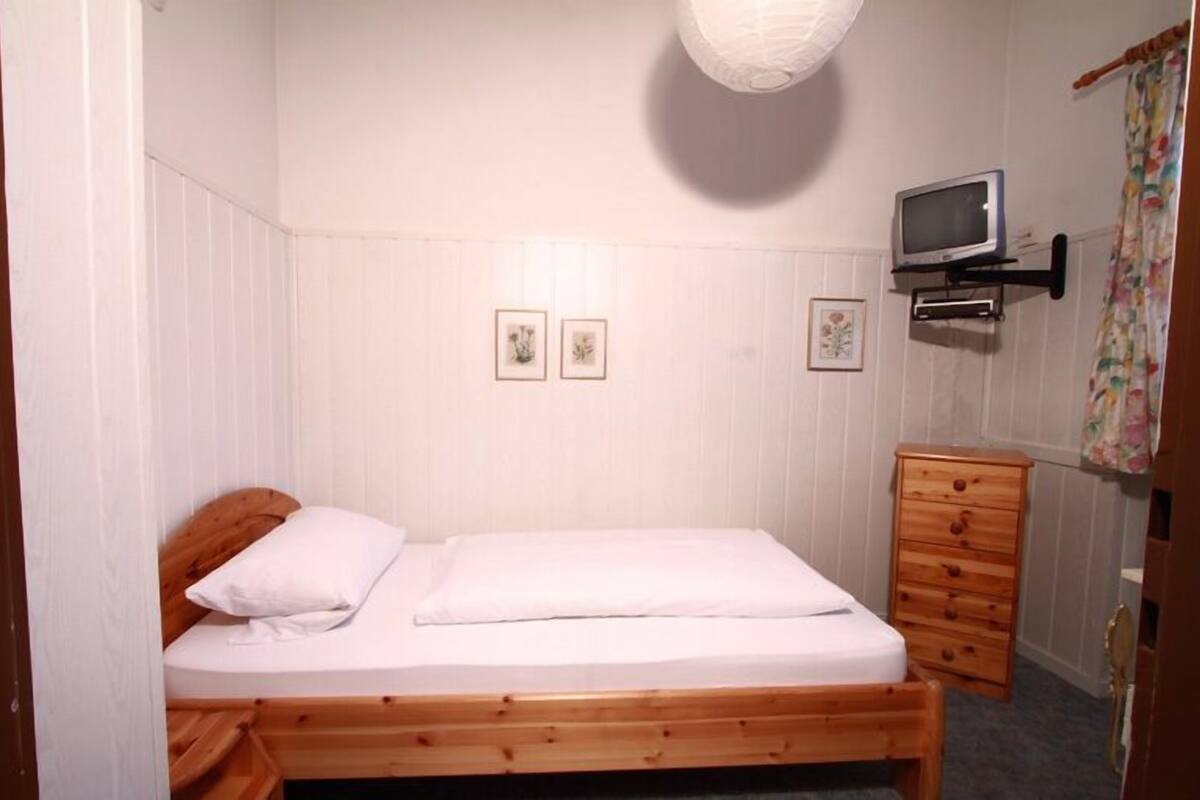 A small, single-bed room with a wooden bed frame, a wooden dresser, and a television mounted on the wall.