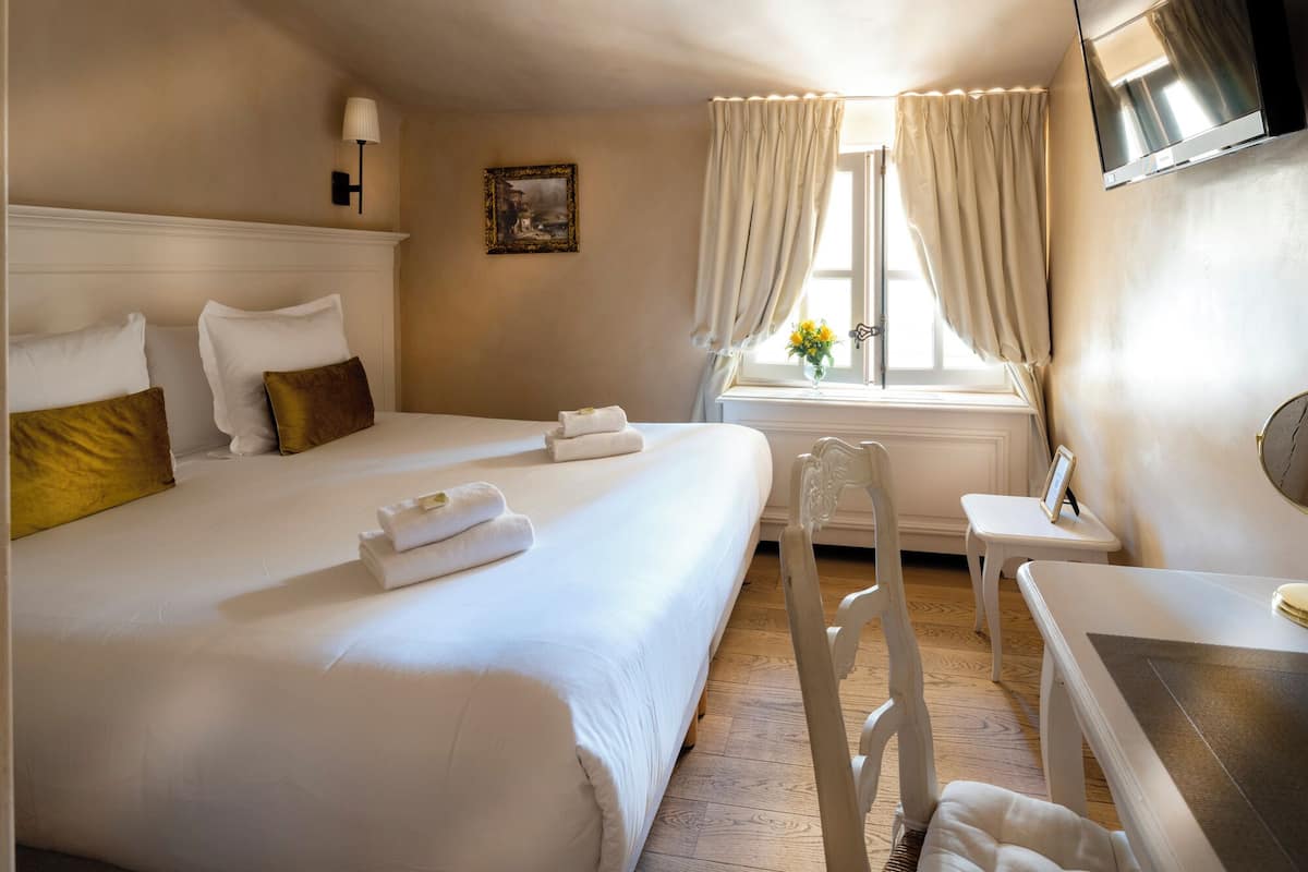 Una habitación de hotel con dos camas, una mesita, una silla, un ventanal con cortinas y un cuadro en la pared.