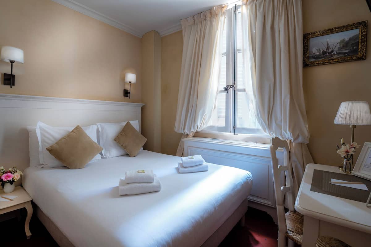 Habitación de hotel con una cama grande, una mesita de noche con lámpara, una ventana con cortinas y un cuadro en la pared.