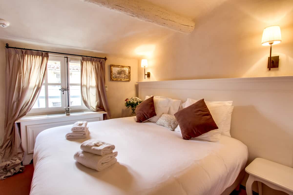 Una cama bien hecha con sábanas blancas y almohadas marrones, una mesita de noche y una ventana con cortinas.