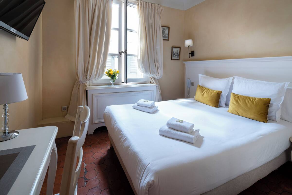 Una habitación de hotel con una cama, un escritorio con lámpara, una ventana con cortinas y un jarrón con flores.
