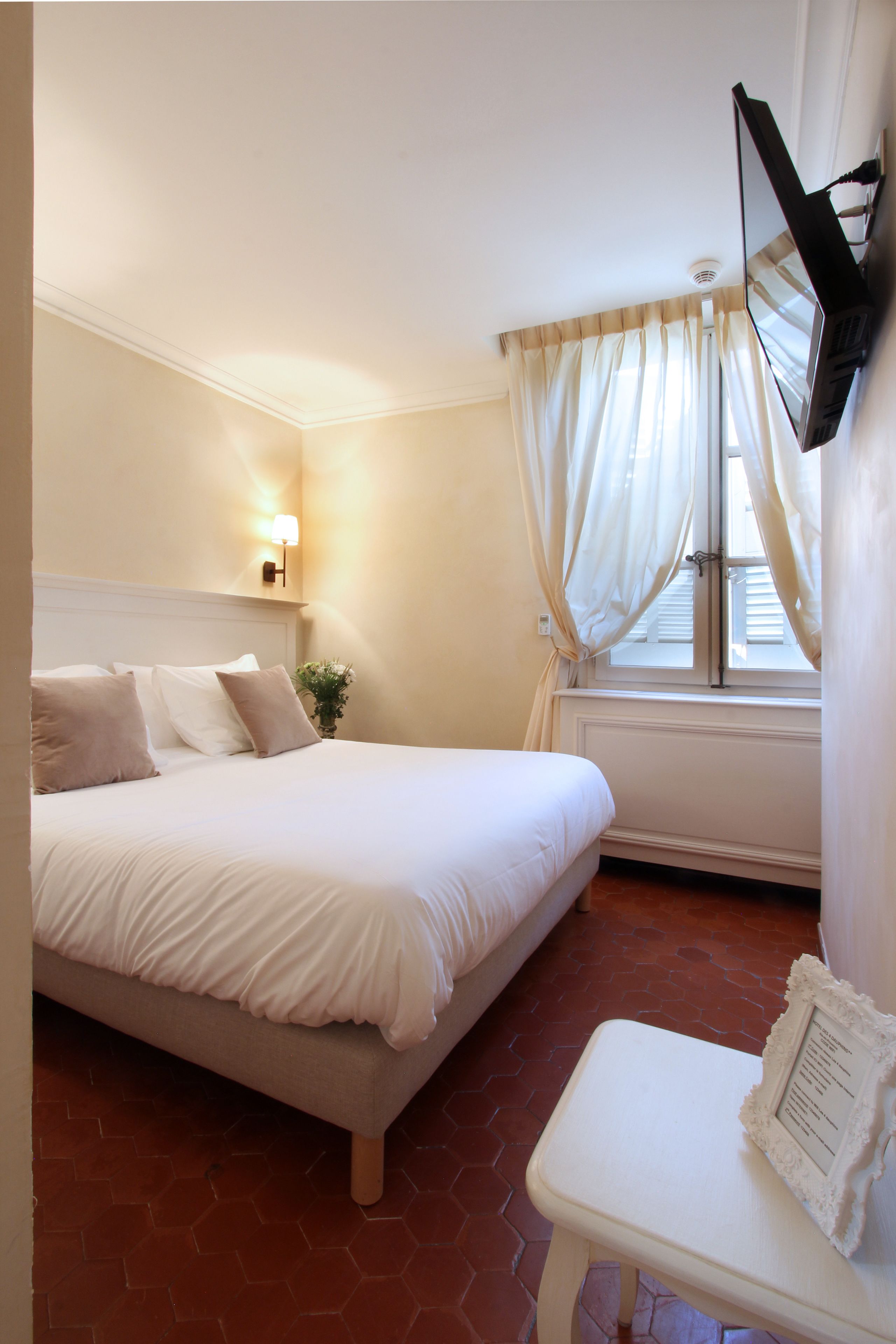 A neatly arranged bedroom with a bed, a bedside table with a picture frame, a wall-mounted TV, and a window with curtains.