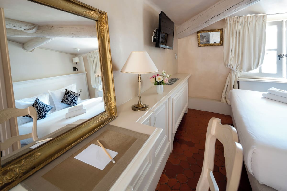 Habitación de hotel con cama, mesita de noche con lámpara y flores, un espejo grande y un televisor montado en la pared.