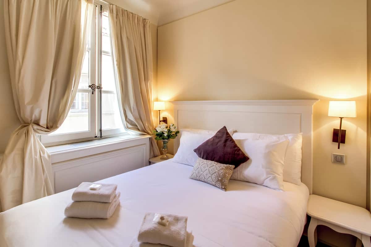 Una cama bien hecha con sábanas blancas y dos toallas dobladas, un almohadón rojo oscuro y un cojín decorativo con un diseño geométrico. Hay dos lámparas de noche a cada lado de la cama, y una ventana con cortinas parcialmente corridas a la izquierda.