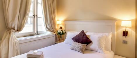 A neatly made bed with white linens and two folded towels, a dark red pillow, and a decorative pillow with a geometric pattern. There are two bedside lamps on either side of the bed, and a window with curtains partially drawn to the left.