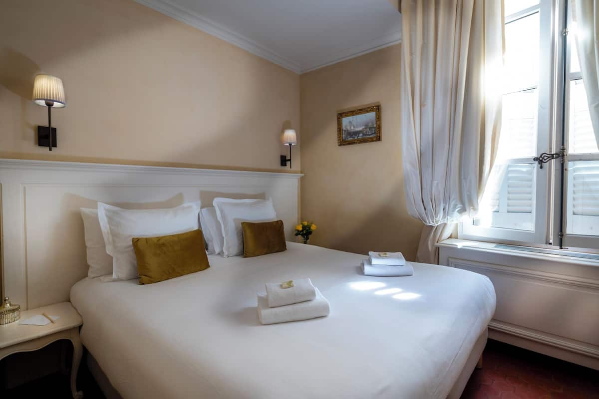 Una cama bien tendida con sábanas blancas y dos almohadas marrones, una mesita de noche con una lámpara, y una ventana con cortinas.